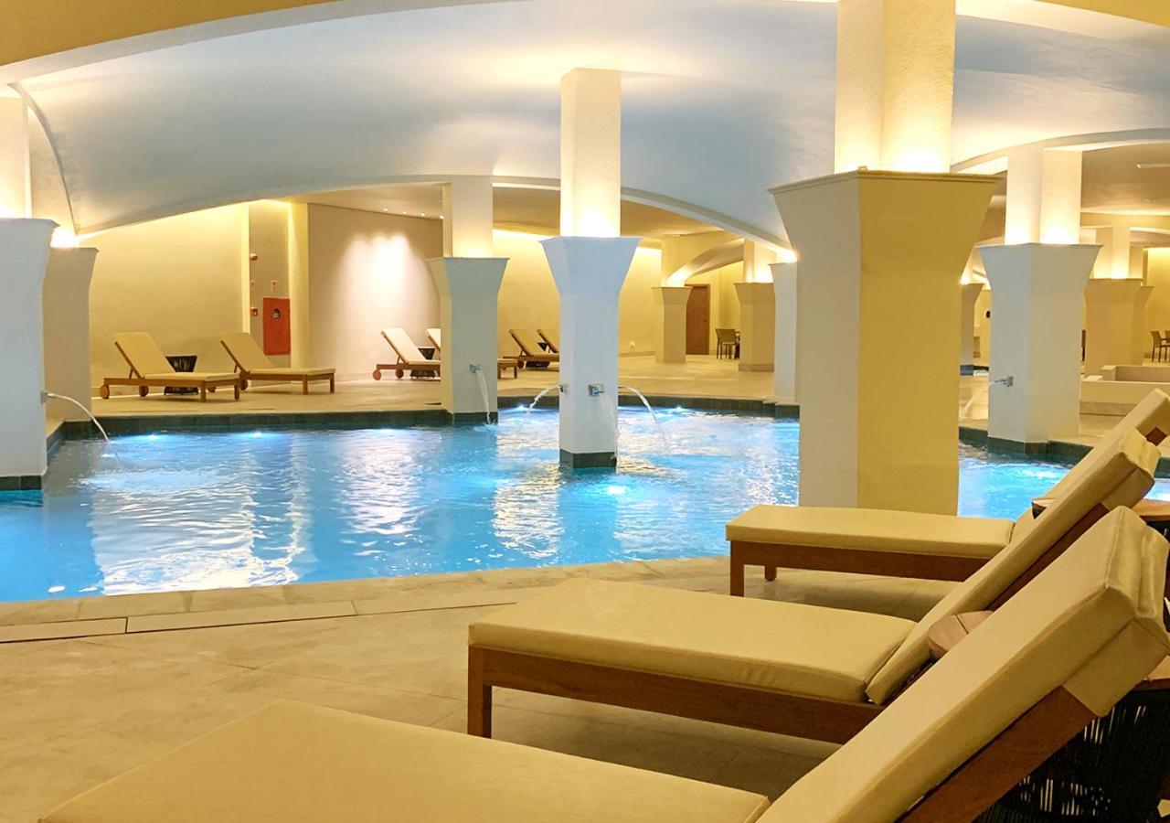 Buona Vitta Gramado Resort & Spa By Gramado Parks ภายนอก รูปภาพ The photo shows an indoor spa or relaxation area with a tranquil atmosphere. There is a large swimming pool with clear blue water, illuminated by soft lighting. Surrounding the pool are several comfortable lounge chairs. The architecture features ele