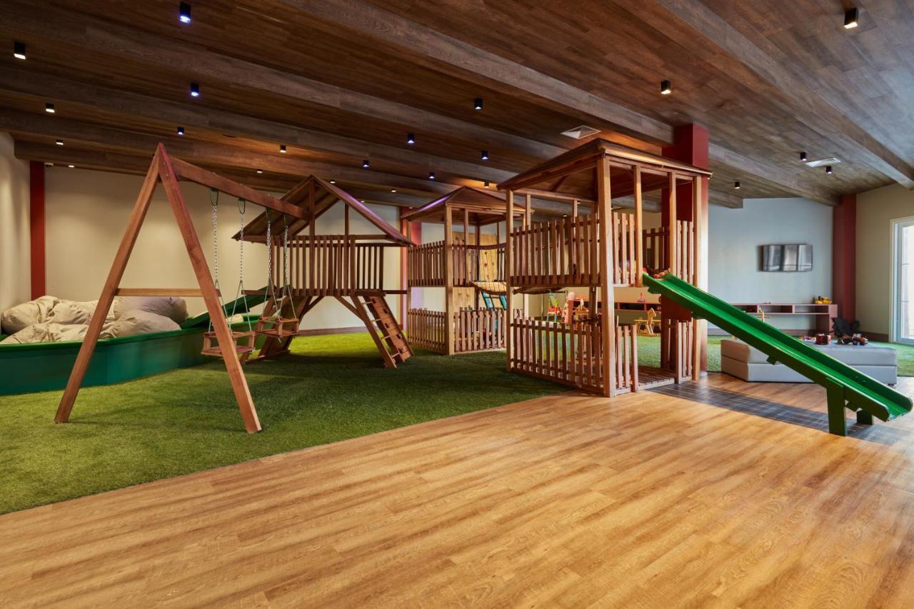 Buona Vitta Gramado Resort & Spa By Gramado Parks ภายนอก รูปภาพ The photo shows an indoor play area designed for children. It features a wooden play structure that includes multiple levels, slides, and a swing set. The floor is covered with wooden planks, and there is a green carpet-like area that adds a soft tou