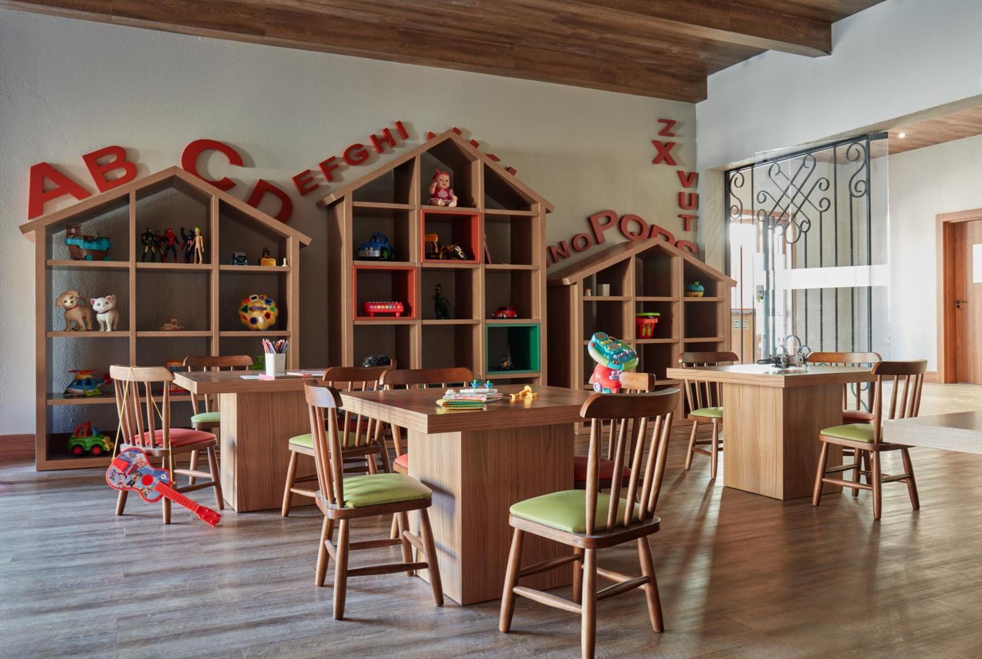 Buona Vitta Gramado Resort & Spa By Gramado Parks ภายนอก รูปภาพ The photo shows a classroom or playroom designed for children. It features wooden furniture, including several tables and chairs with green cushions. In the background, there are large, open shelving units shaped like houses, displaying various toys 