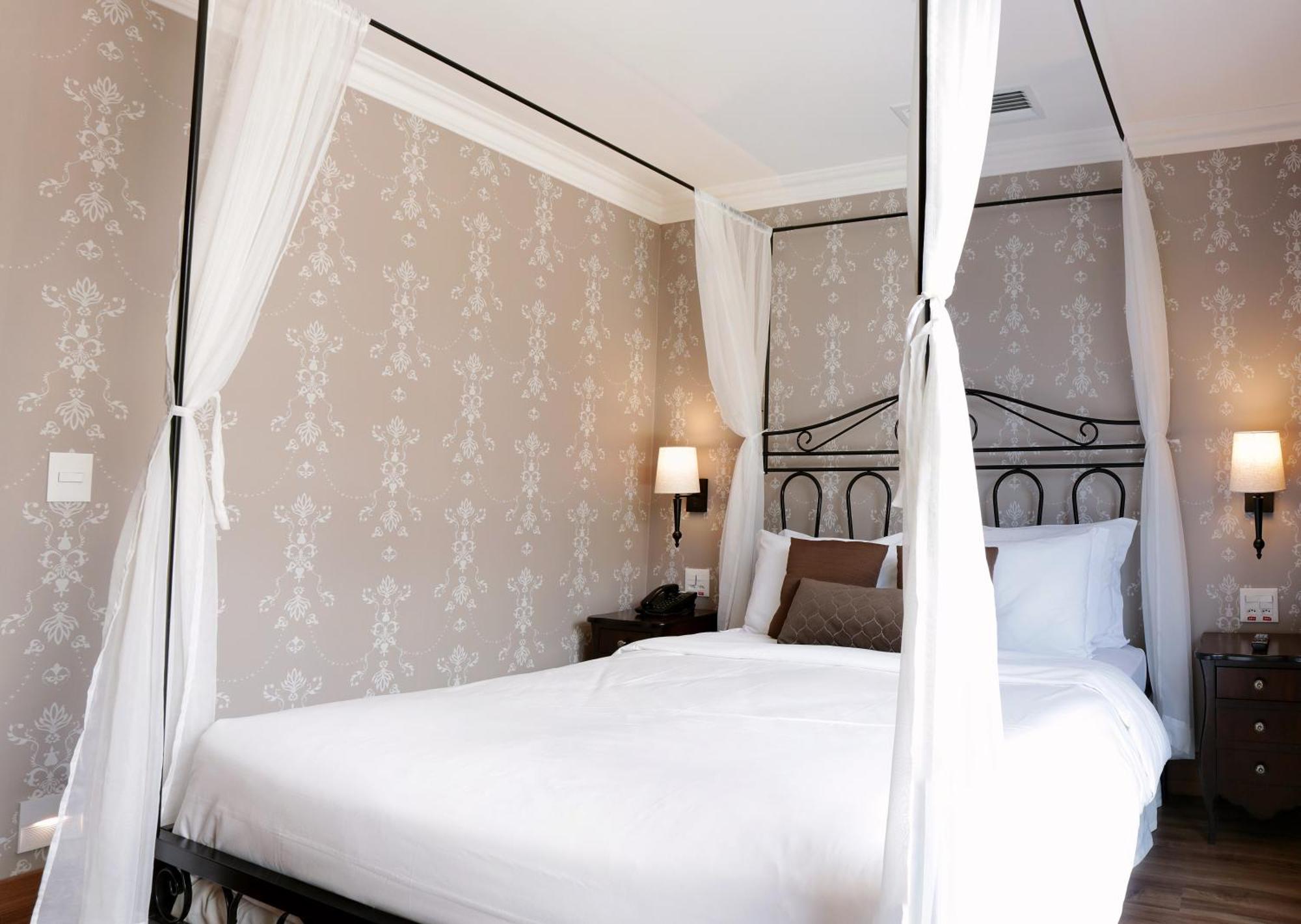 Buona Vitta Gramado Resort & Spa By Gramado Parks ภายนอก รูปภาพ The photo shows a elegantly decorated bedroom featuring a four-poster bed with white drapes. The bed is made with white linens and has some brown accent pillows. The walls are adorned with a subtle floral pattern, and there are two bedside lamps prov