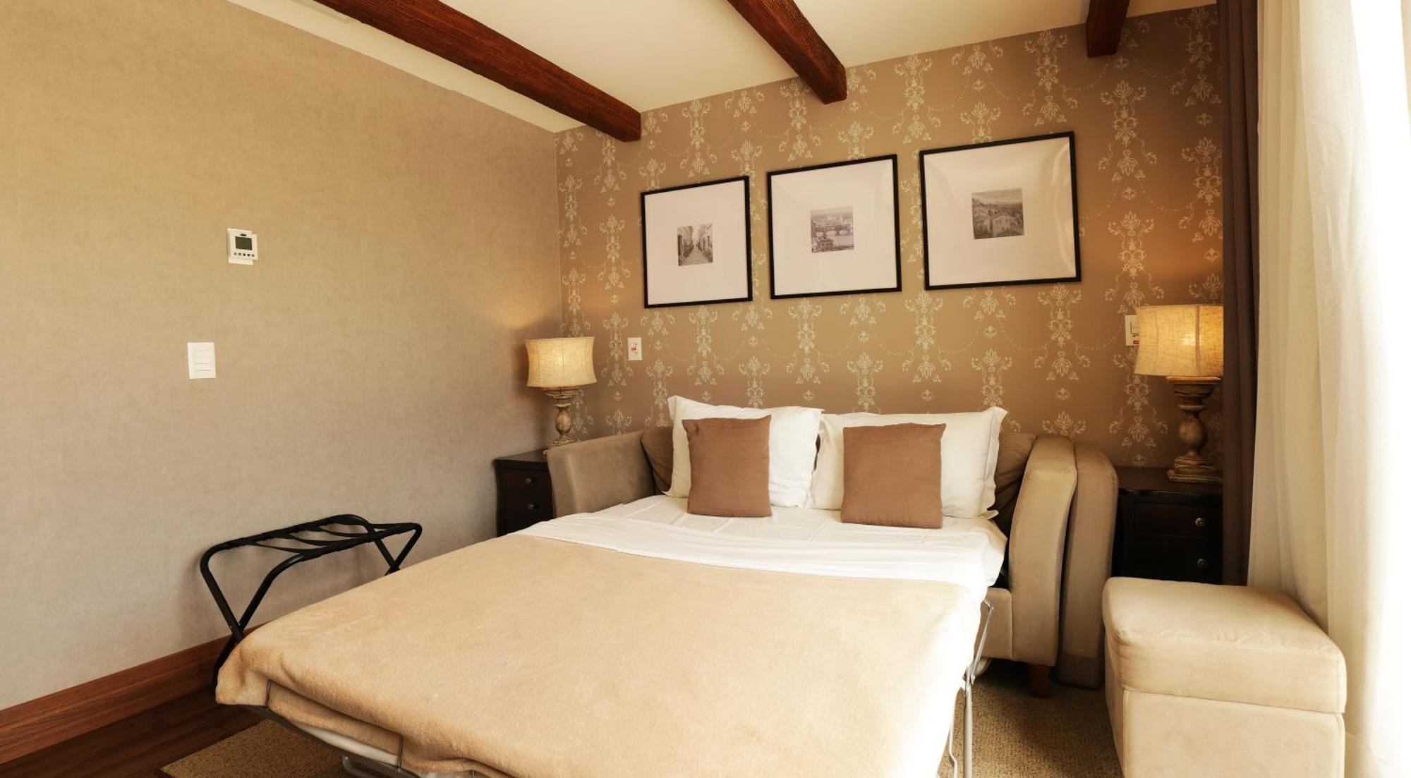 Buona Vitta Gramado Resort & Spa By Gramado Parks ภายนอก รูปภาพ The photo shows a cozy bedroom with a neutral color palette. There is a bed with white linens and two brown decorative pillows. On either side of the bed, there are bedside tables with lamps. The walls feature a subtle floral pattern, and three frame