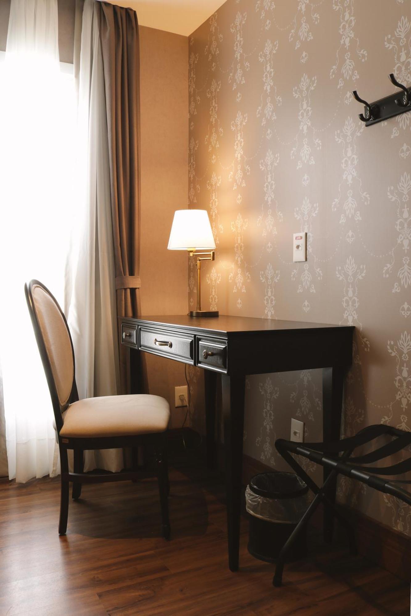 Buona Vitta Gramado Resort & Spa By Gramado Parks ภายนอก รูปภาพ The photo shows a small desk set against a wall with a decorative pattern. The desk is black with a few drawers and has a small lamp on it. Next to the desk, there is a chair with a light-colored cushion. Soft light is coming through a window, castin